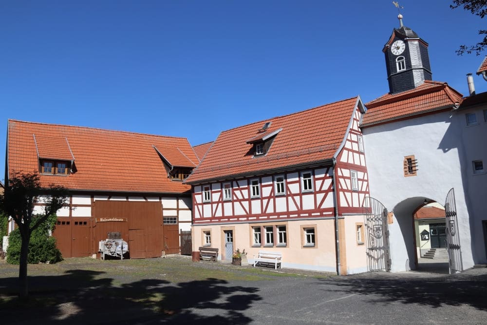 In Kaltennordheim steht unter strahlend blauem Himmel ein traditionelles deutsches Fachwerkhaus mit rotem Ziegeldach und einem Uhrturm neben einem großen Holztor. Im Innenhof sind Bänke und ein Karren zu sehen.