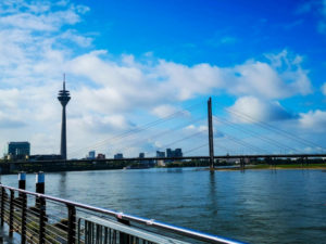 Eine atemberaubende Flussansicht von Düsseldorf, Deutschland, mit dem Rheinturm und der Rheinknie-Brücke unter einem blauen Himmel mit vereinzelten Wolken. Moderne Gebäude säumen das andere Ufer und zeigen das pulsierende Stadtbild von Düsseldorf.