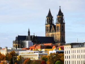 Ein Blick auf eine Kathedrale im gotischen Stil mit zwei hohen Türmen, die an die Architektur Magdeburgs erinnern und sich über eine Stadtlandschaft erheben. Das Gebäude ist von verschiedenen städtischen Strukturen und leuchtenden Herbstbäumen unter einem klaren Himmel umgeben.