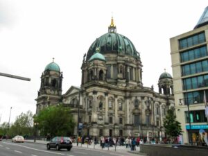 Das Bild zeigt den Berliner Dom, ein großes historisches Gebäude mit einem grünen Kuppeldach, kunstvoller Architektur und Zwillingstürmen im Herzen Berlins. Auf der Straße vor der Kathedrale sind Menschen und Autos zu sehen.